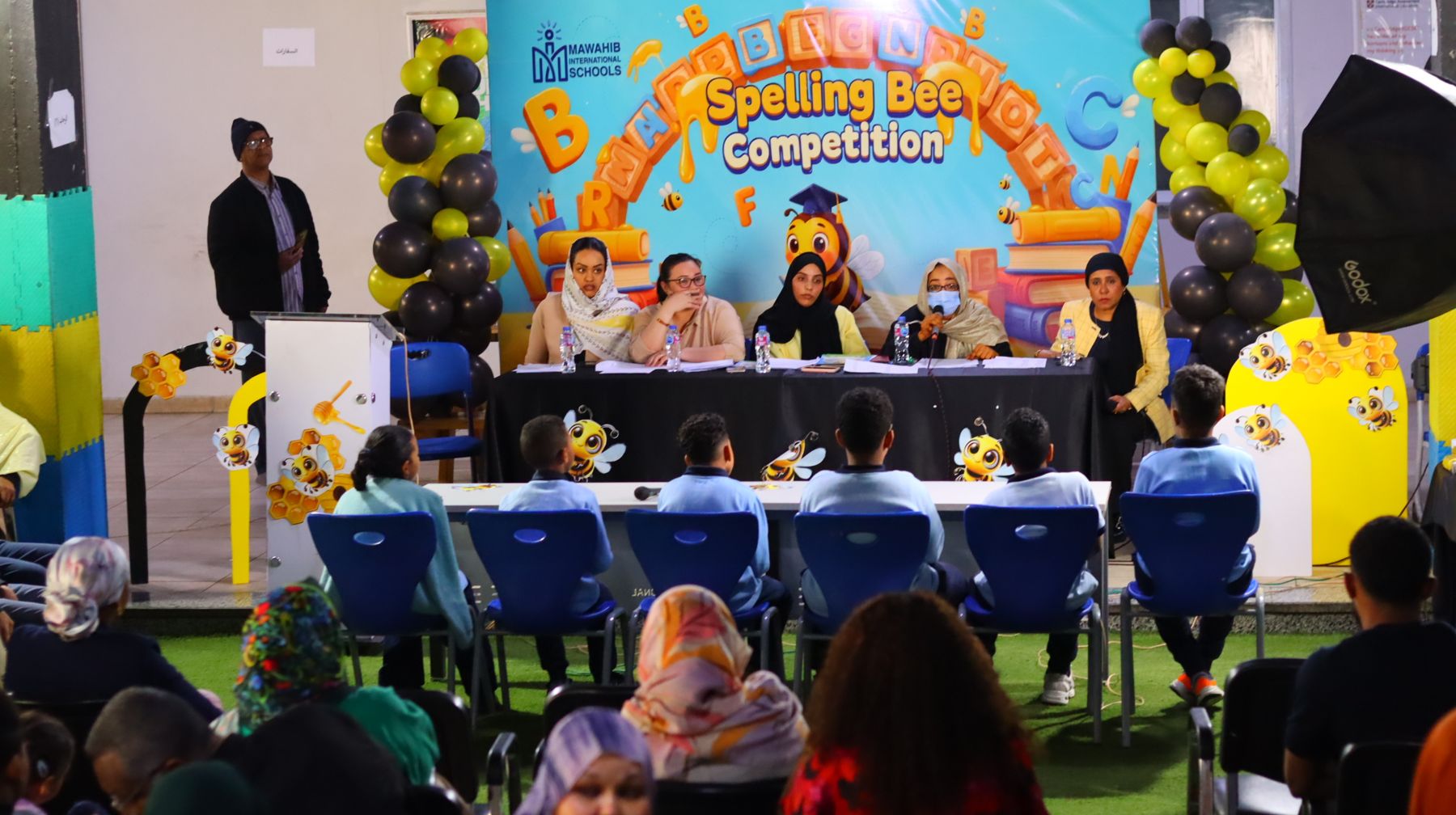 Judges and students on stage at the Spelling Bee competition, with families watching
